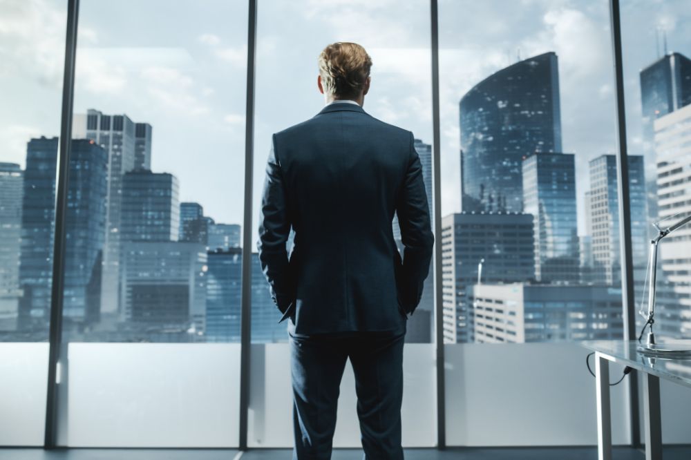 světlovlasý muž v tmavém obleku stojící zády před prosklenou stěnou s výhledem na mrakodrapy